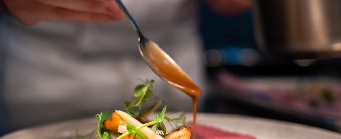 A hand holding a spoon which is drizzling gravy onto a dish